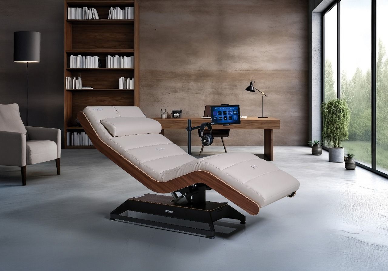 Modern clinic room with an adjustable treatment bed, desk, bookshelf, and large windows overlooking greenery