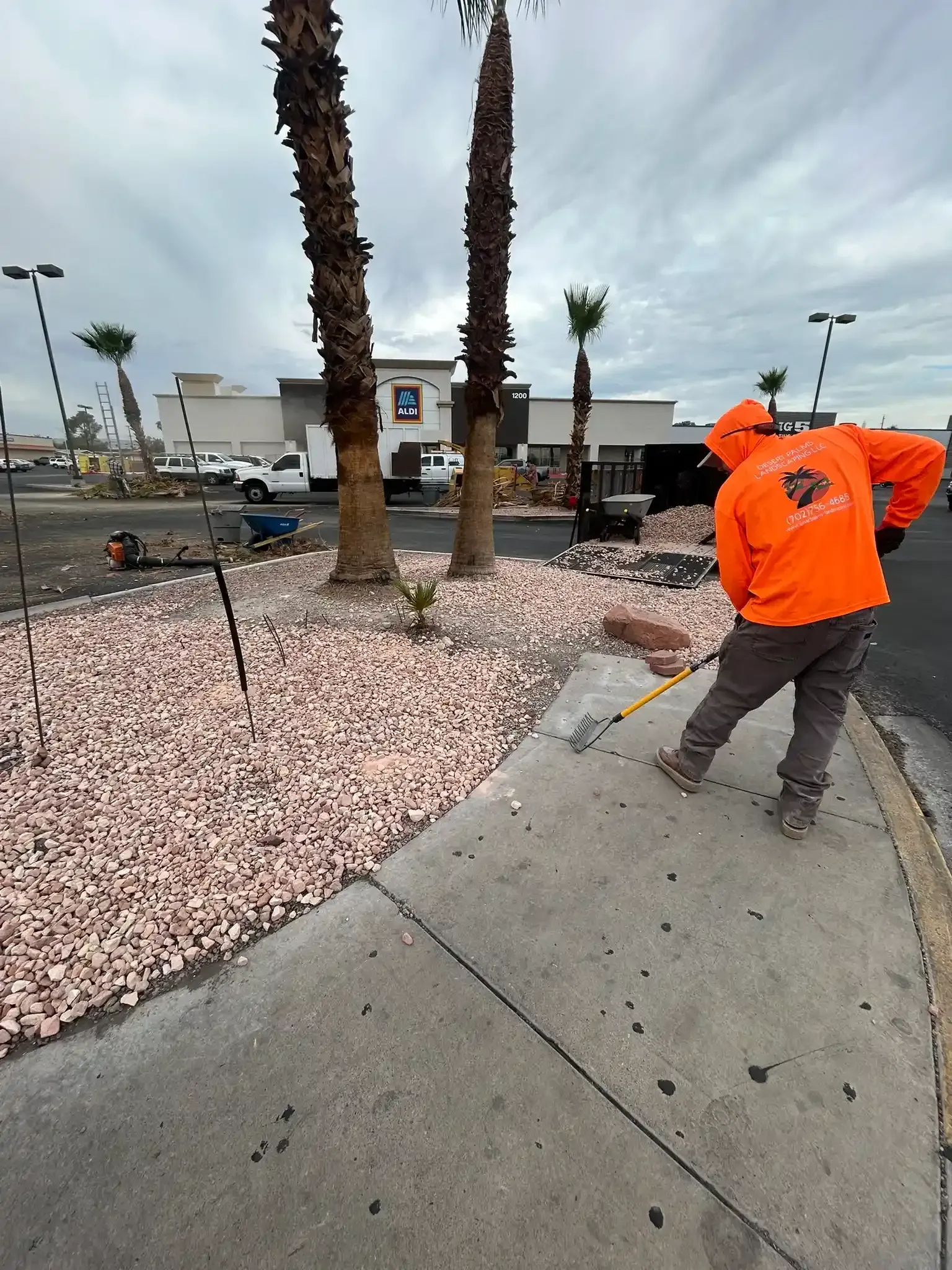 Sidewalk beside parked vehicles and hedges, with a worker in a neon vest under a sunny sky.