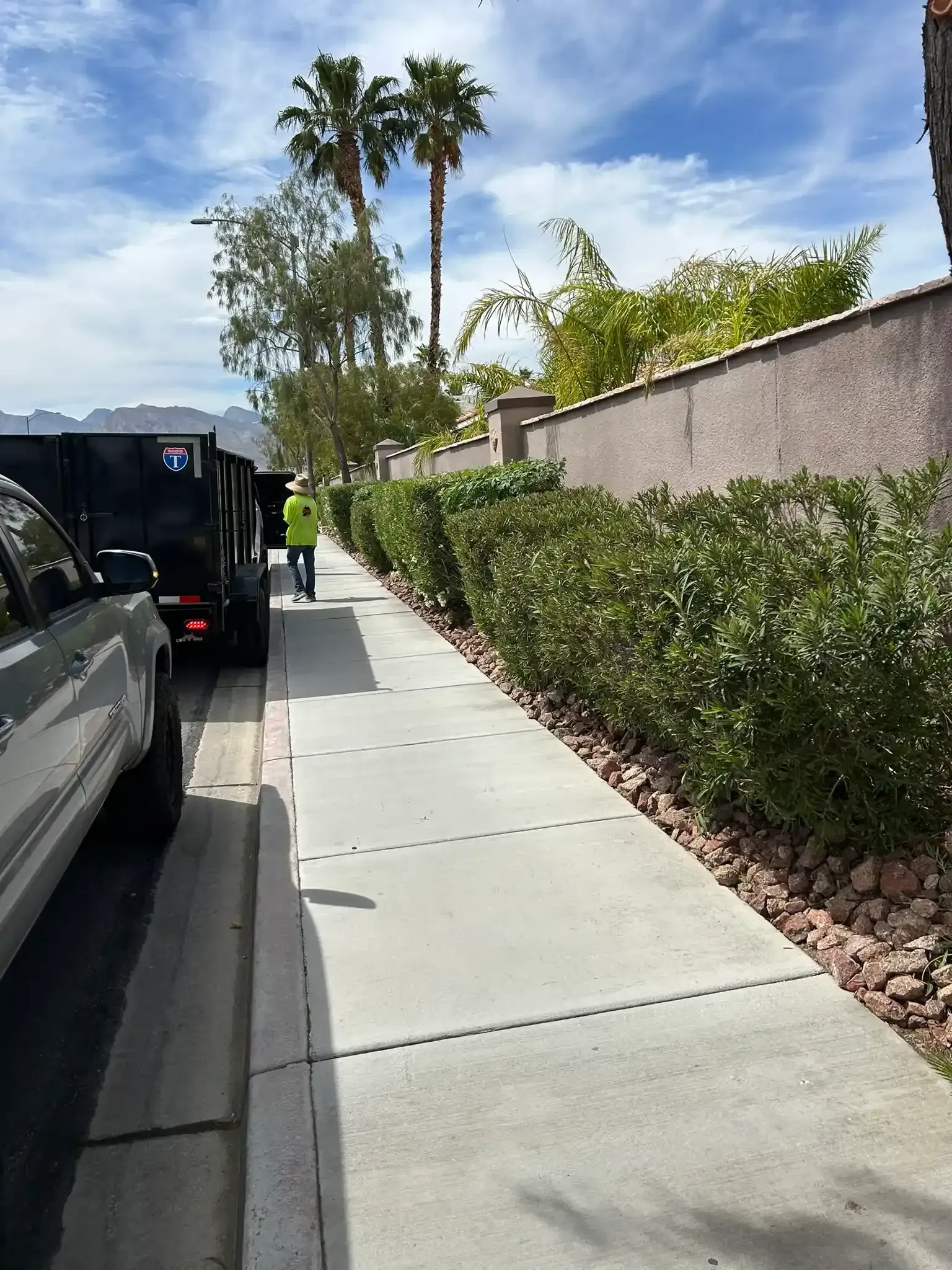 Sidewalk beside parked vehicles and hedges, with a worker in a neon vest under a sunny sky.