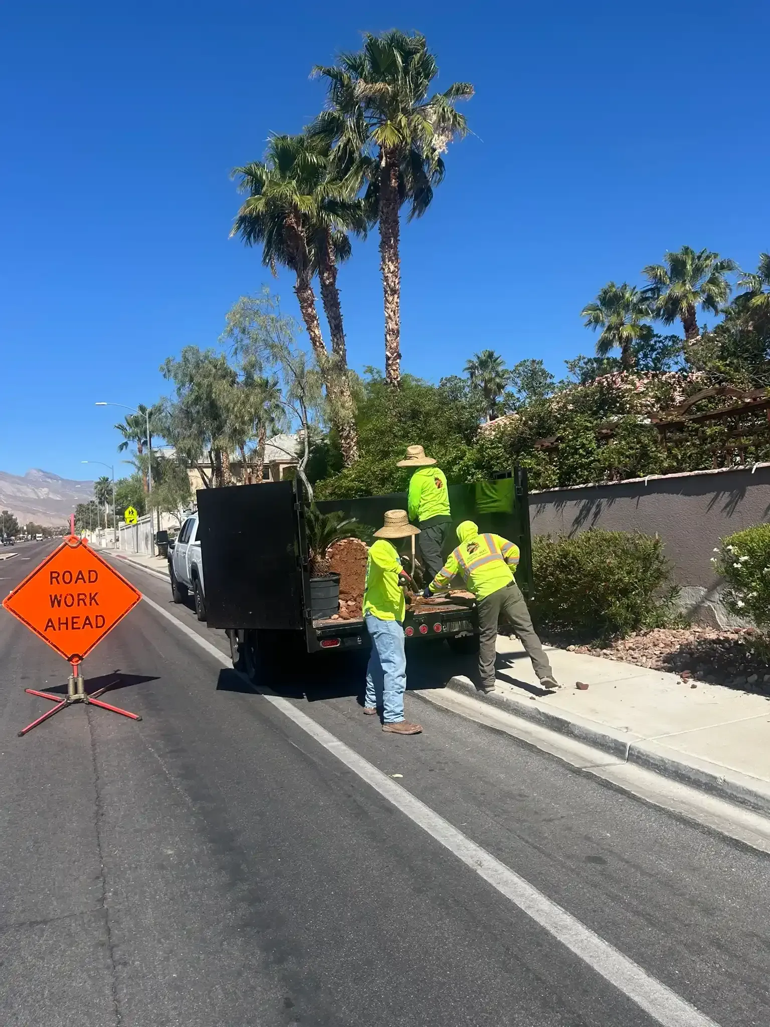 Workers loading a truck on a road beside an orange construction sign and palm trees.