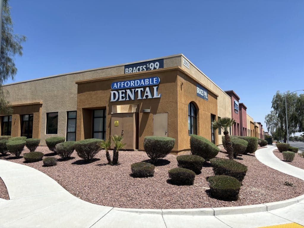 Affordable Dental office building with a blue sign, brown exterior, and landscaping on a sunny day.
