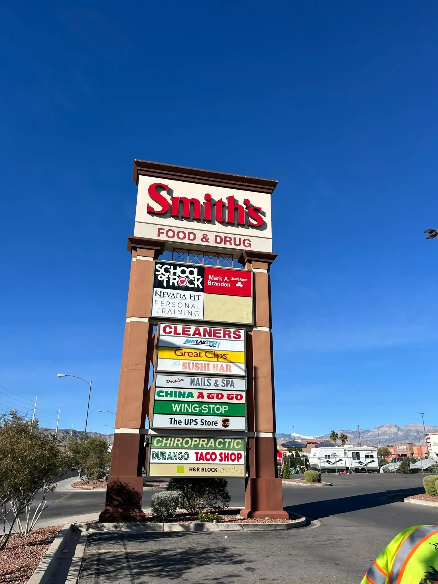 Tall roadside shopping center sign under a blue sky, with multiple store logos listed on panels.
