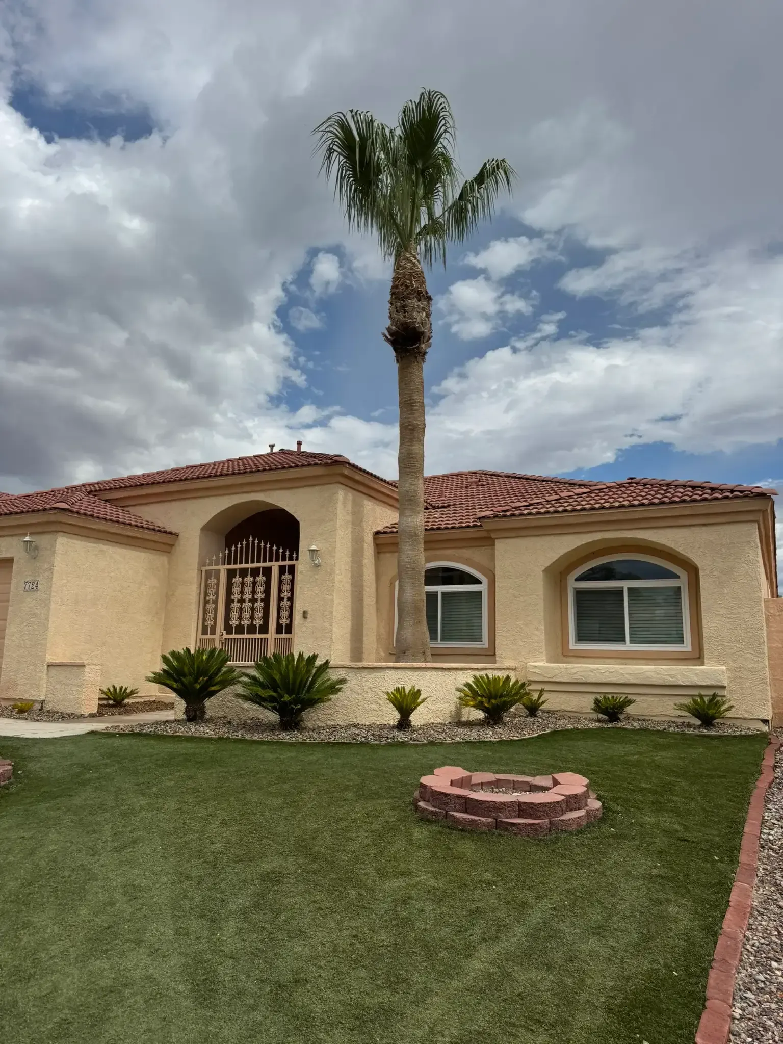 Single-story beige house with red tile roof, tall palm tree, and manicured lawn under a cloudy sky