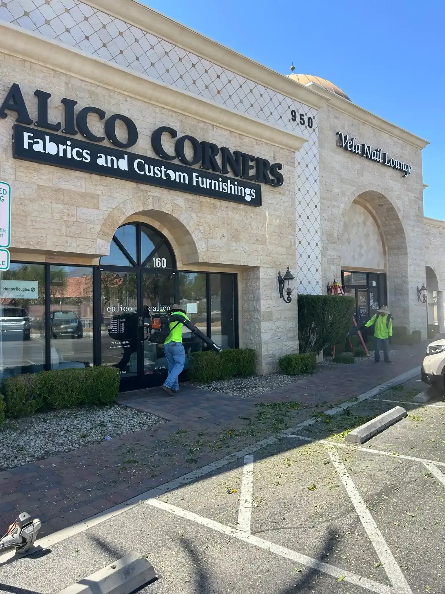Two workers at Alco Corners storefront, one kneeling by the entrance on a sunny day.