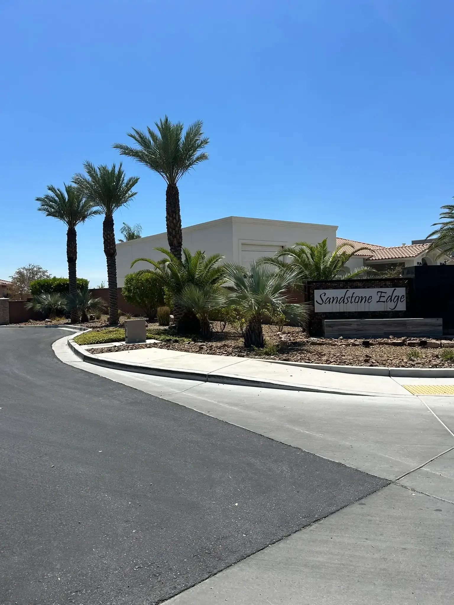 Palm-lined driveway entrance with a beige building, blue sky, and a sign along a curved asphalt road
