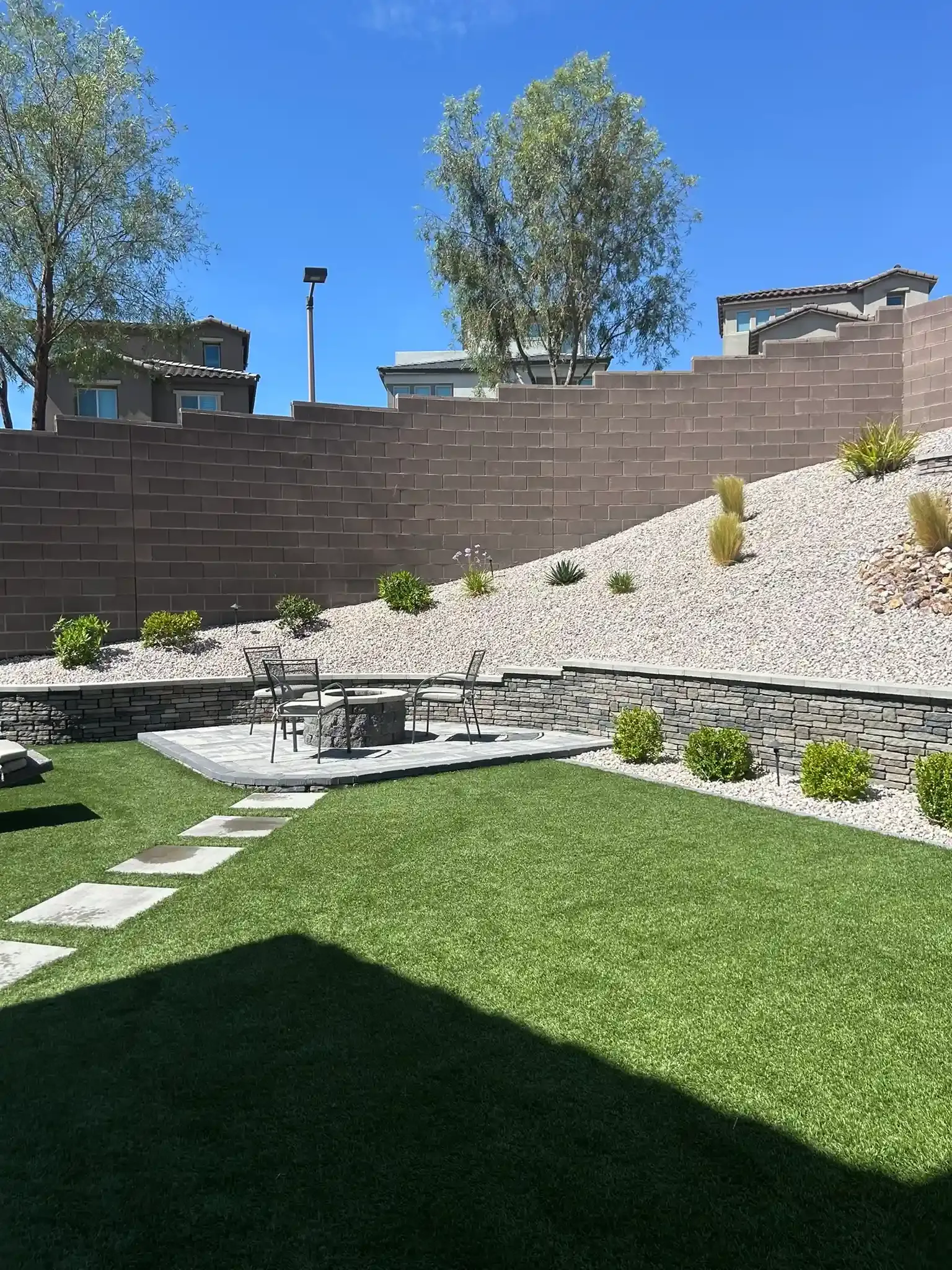 Backyard patio with green lawn, stone steps, and a rocky slope bordered by a brick wall and trees.