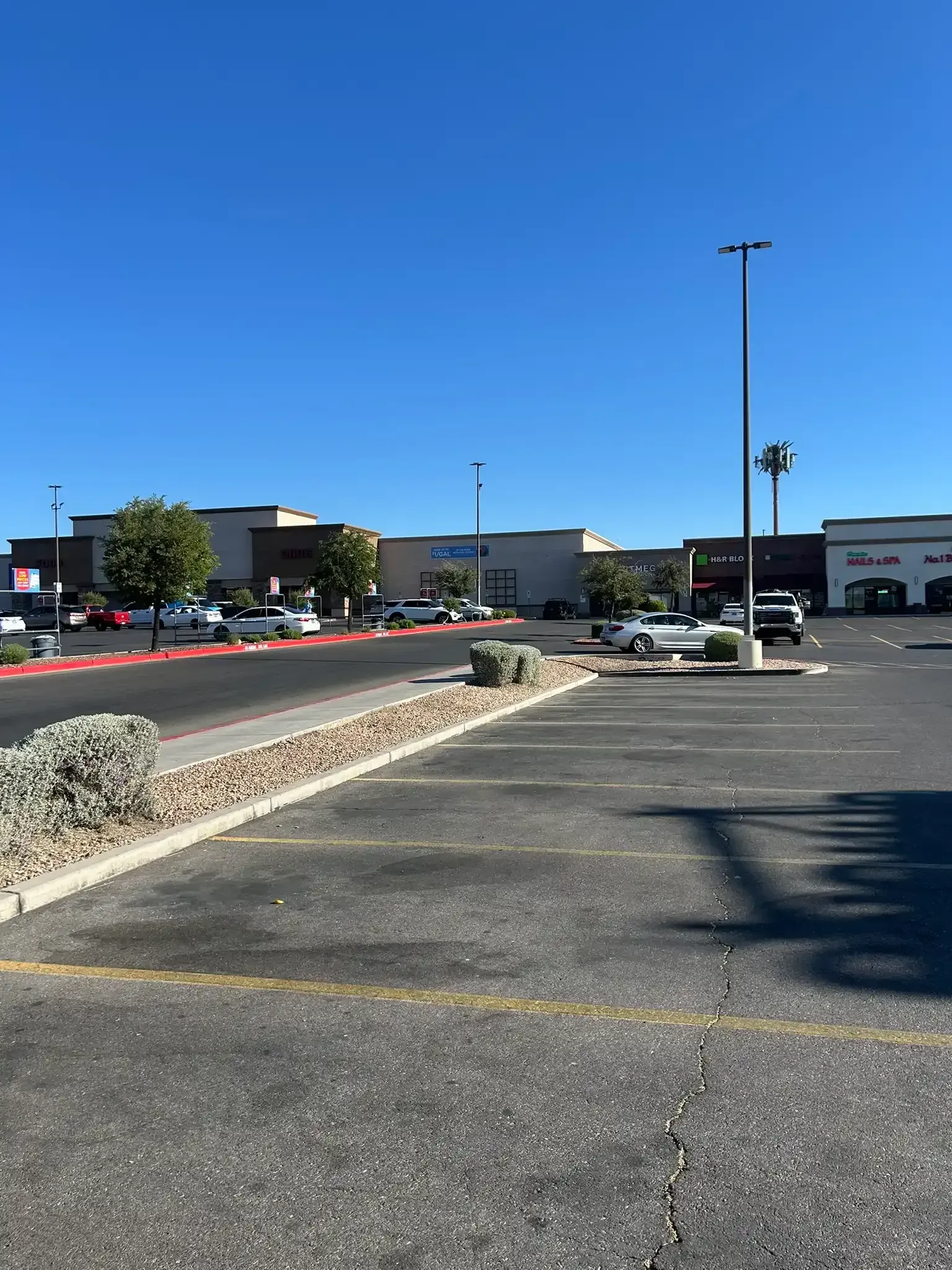 Empty parking lot with cracked asphalt, red curb, and storefronts under a clear blue sky.