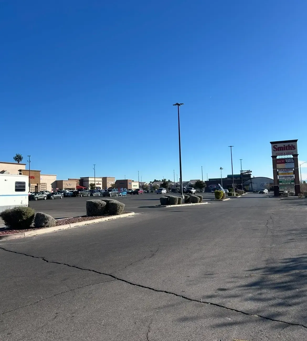 Wide shopping center parking lot under a clear blue sky, with storefronts and a tall sign in the distance.