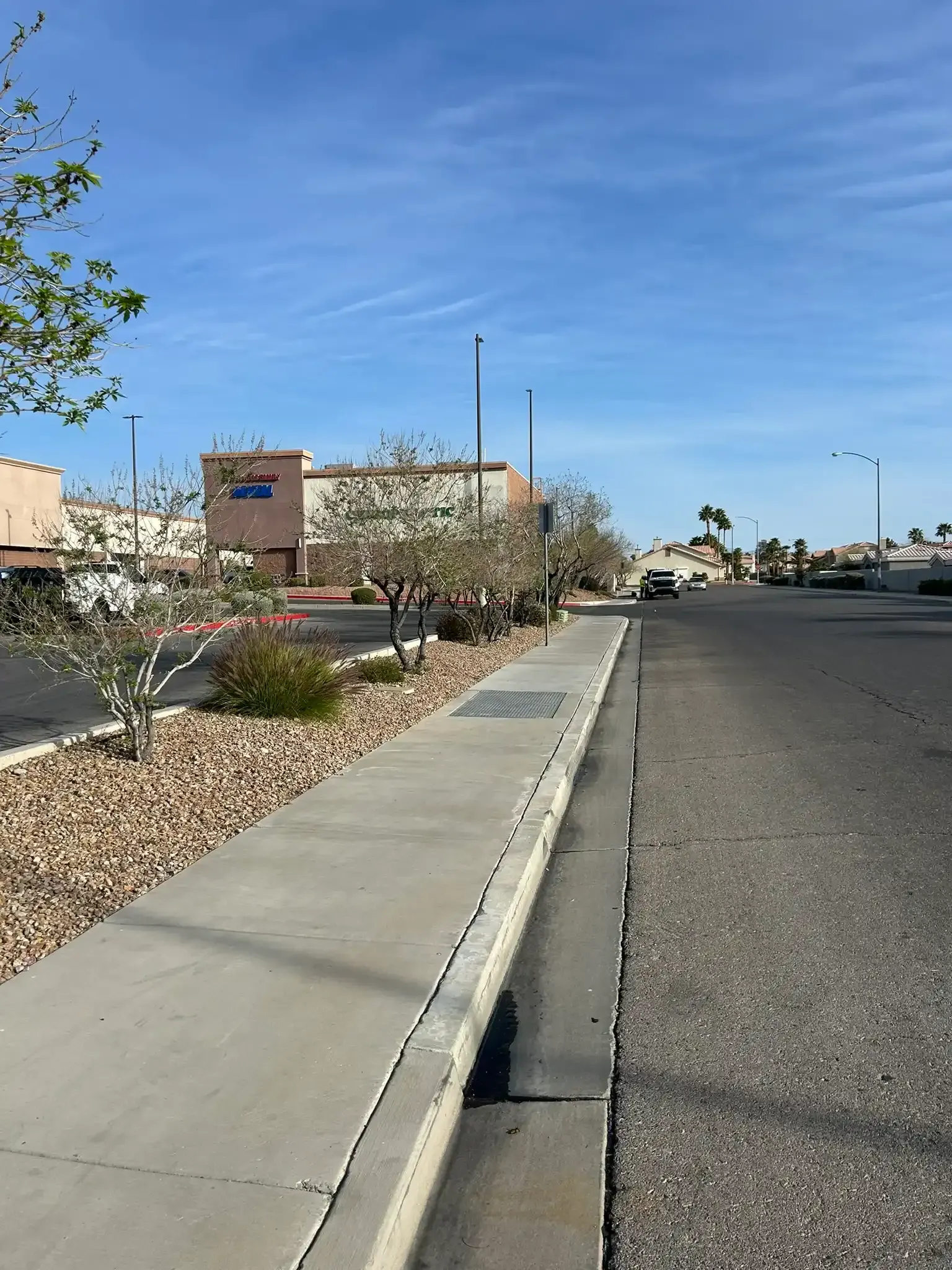 Sidewalk beside a desert parking lot with buildings, shrubs, and a clear blue sky.