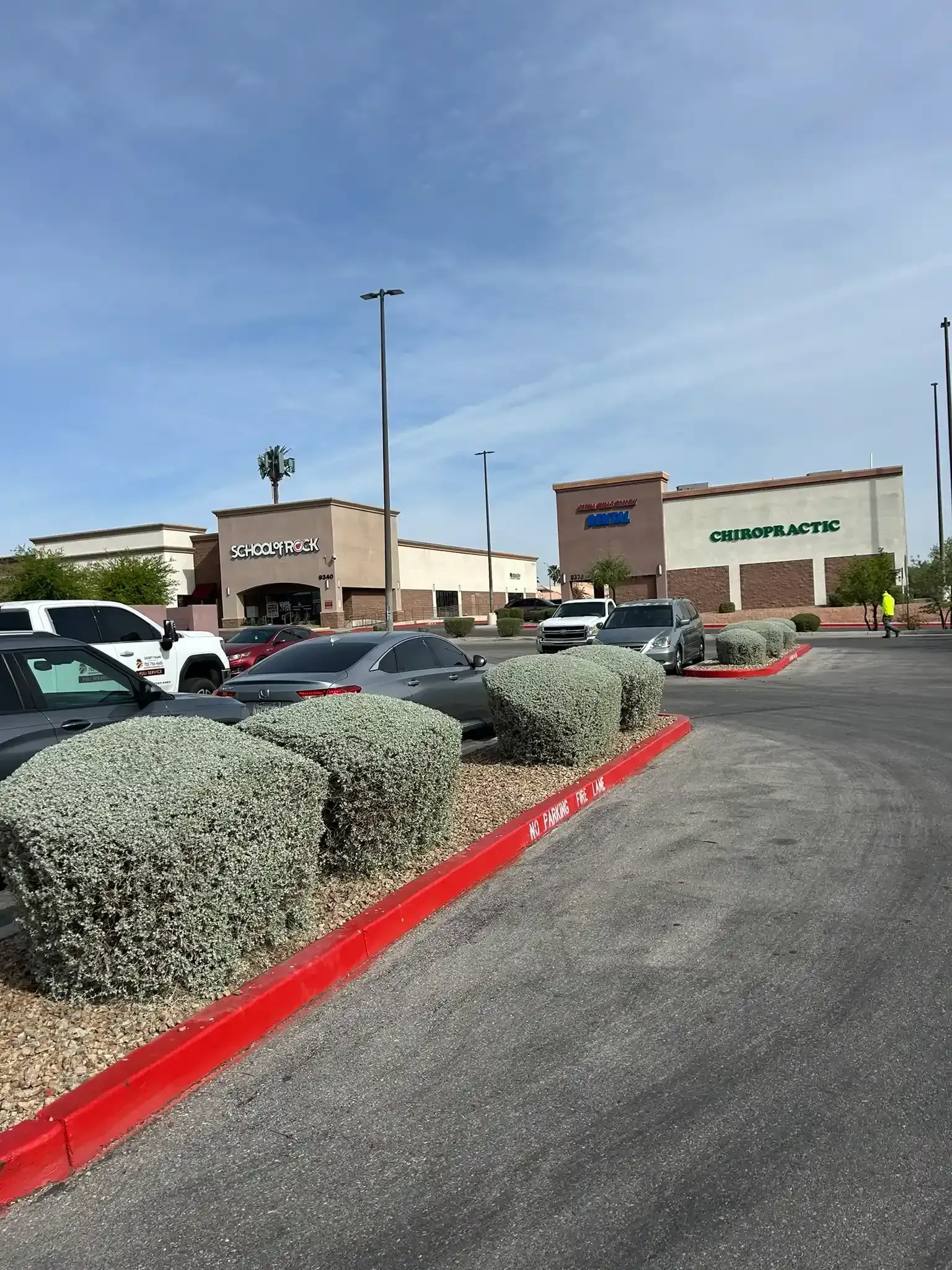 Parking lot with parked cars, red curb, shrubs, and storefront buildings under a clear blue sky