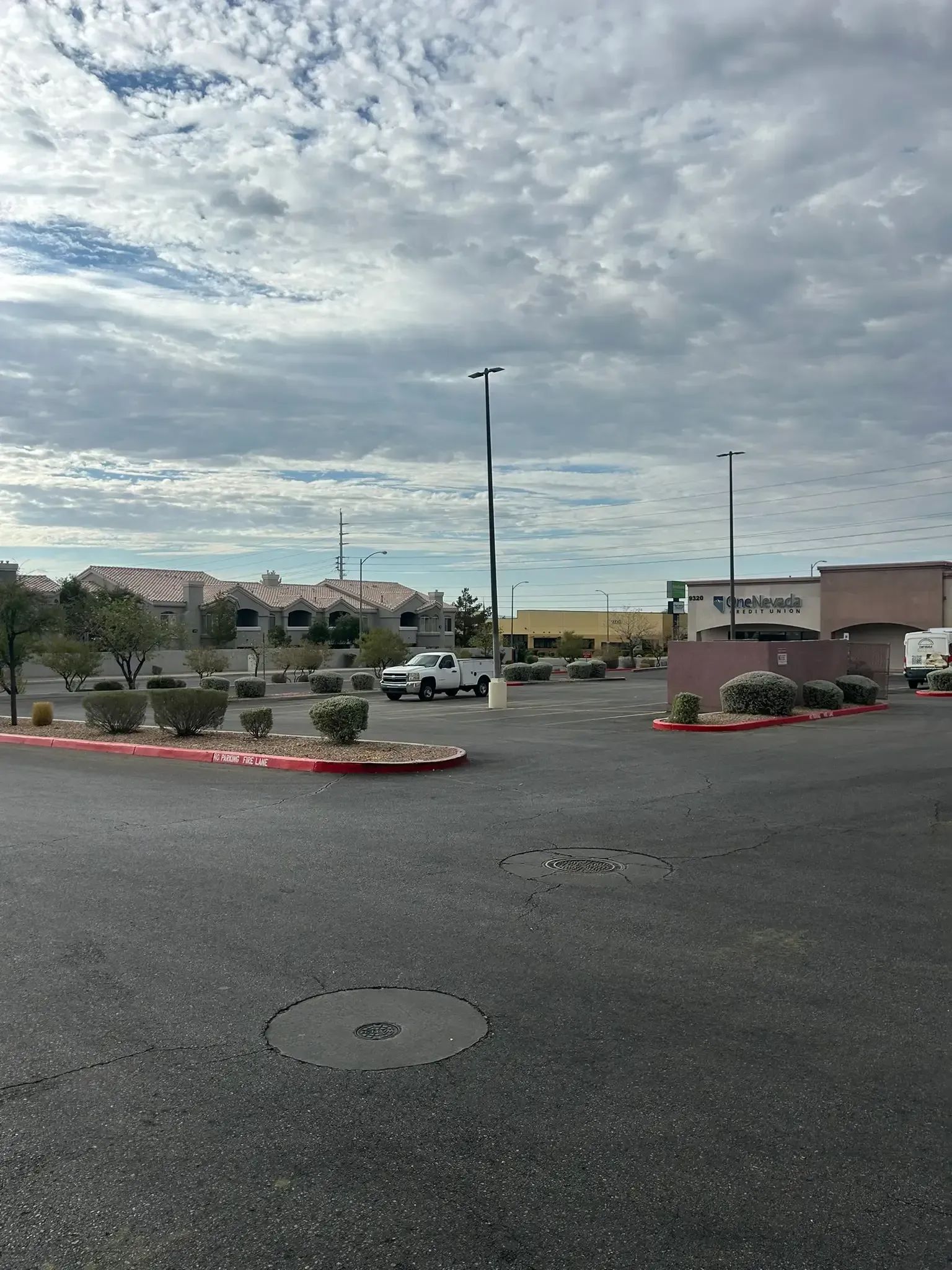 Wide parking lot with red curbs, a few cars, and low buildings under a cloudy sky