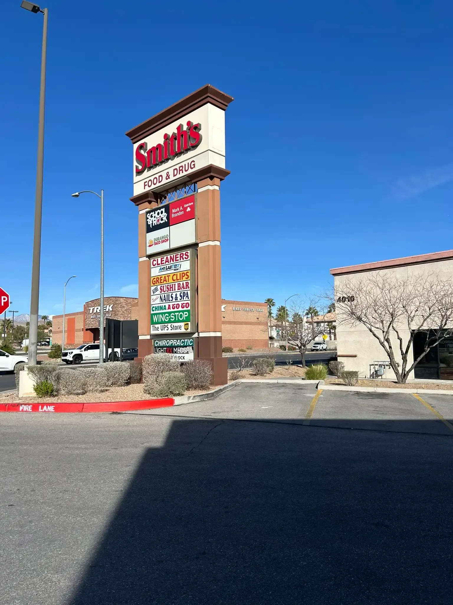 Shopping center sign by a parking lot under a clear blue sky, with storefronts and a brick building nearby.