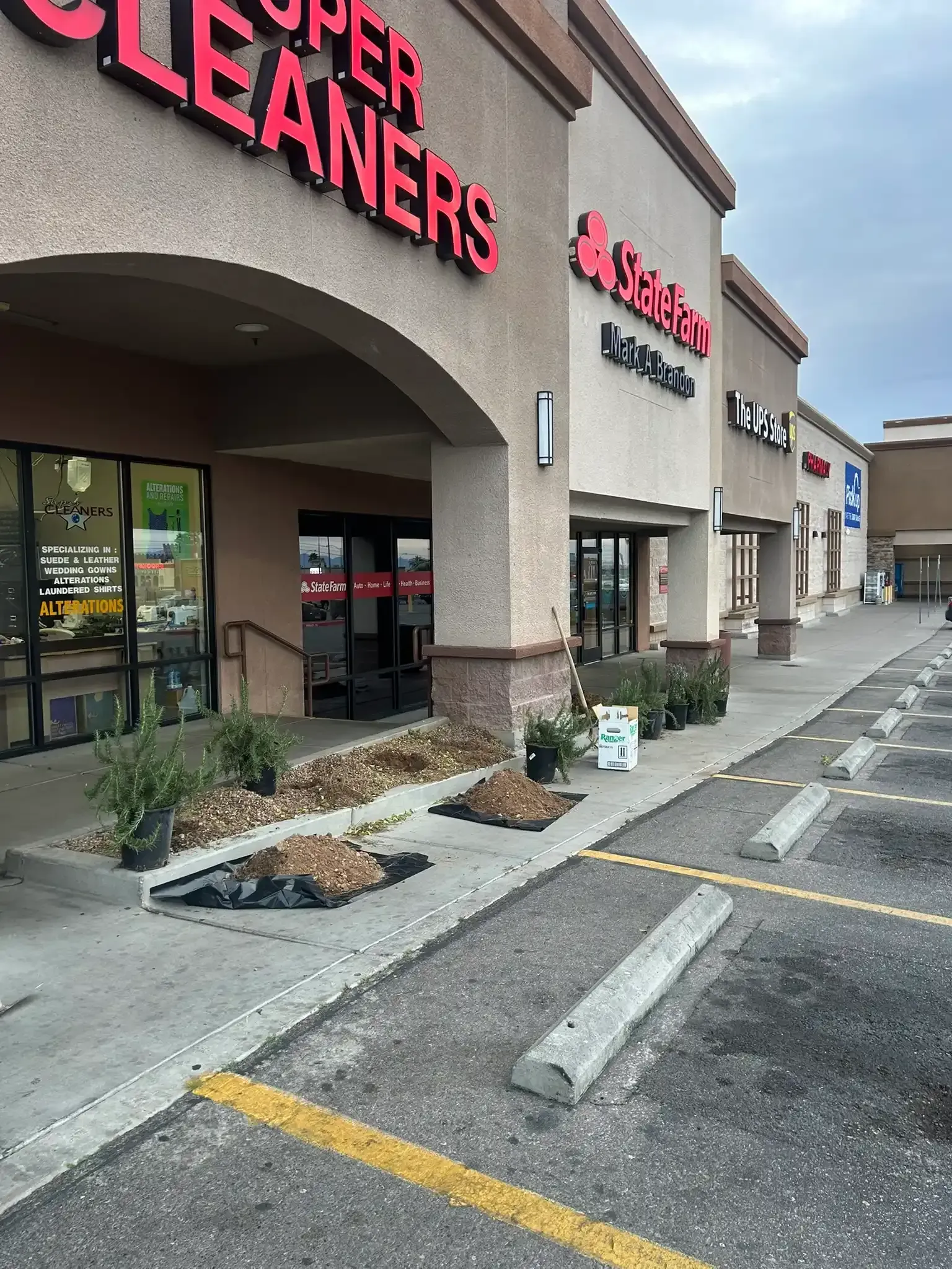 Strip mall storefronts with sidewalk planters and parking spaces, including a Super Cleaners sign.