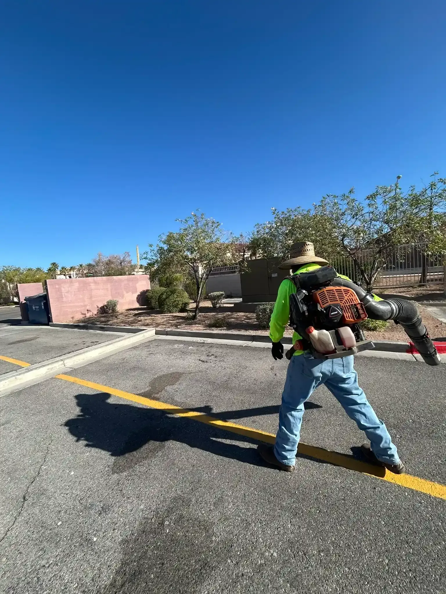 Worker with backpack blower clearing debris on a sunny parking lot road near a pink wall.