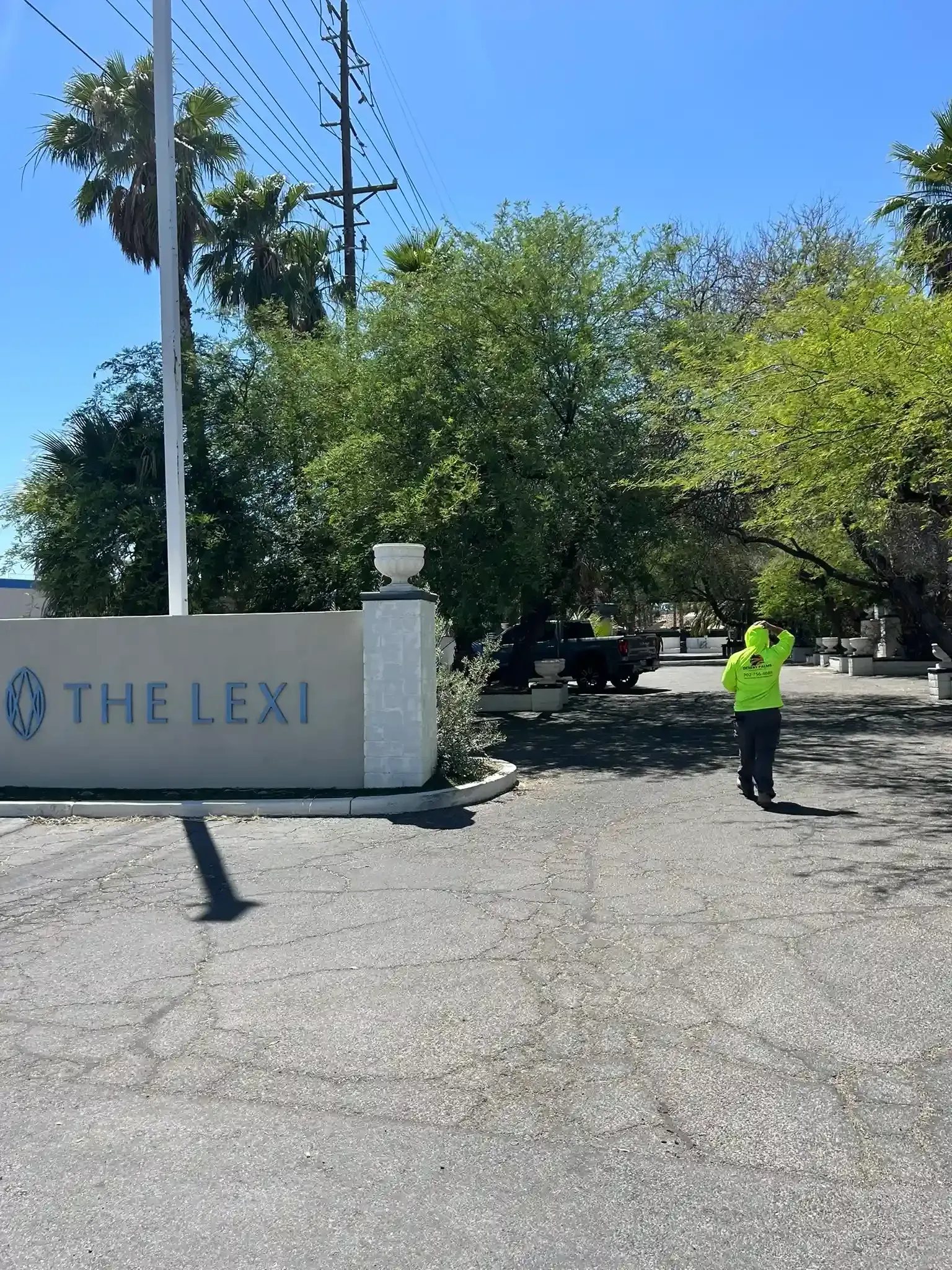 Entrance sign for The Lexi with a person in a neon shirt standing on a gravel driveway under trees