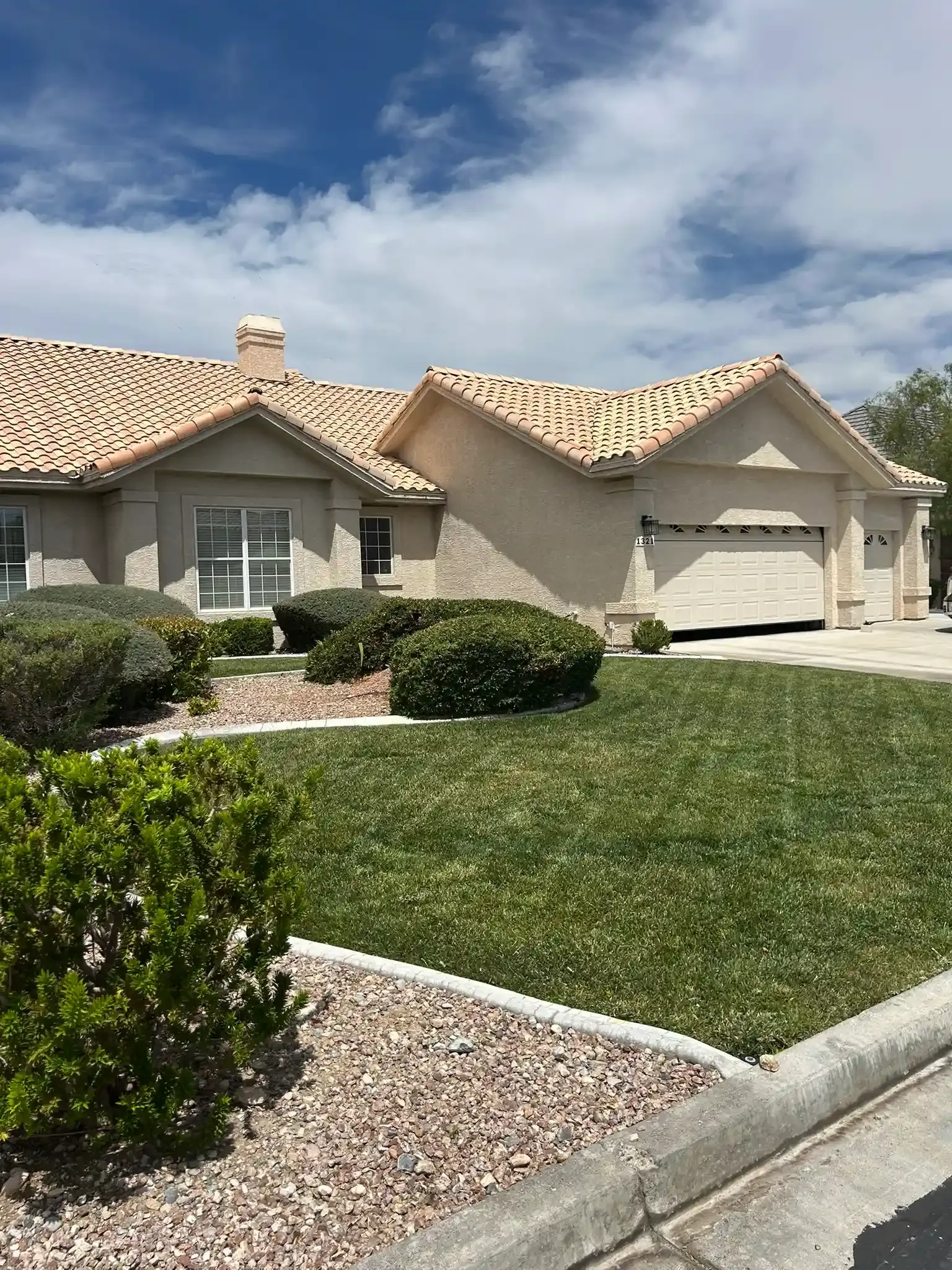 Single-story beige house with a tiled roof, green lawn, and shrubs under a blue sky.