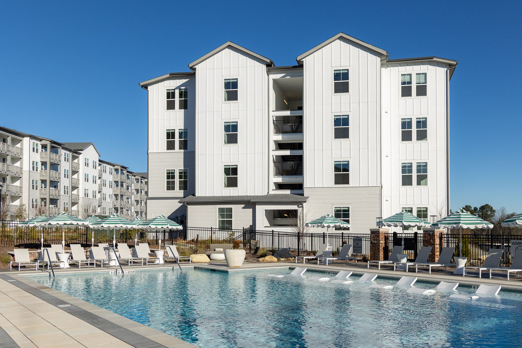 Swimming pool in courtyard of white apartment buildings under a blue sky. Lounge chairs and umbrellas.