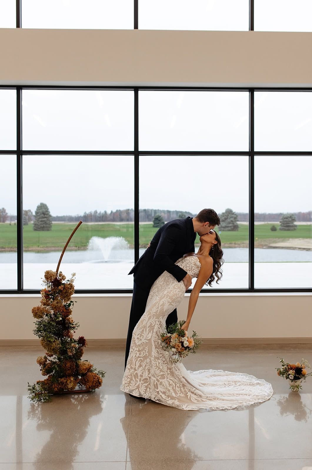 Bride and groom kissing beside a floral arrangement in a bright hall with large window panes