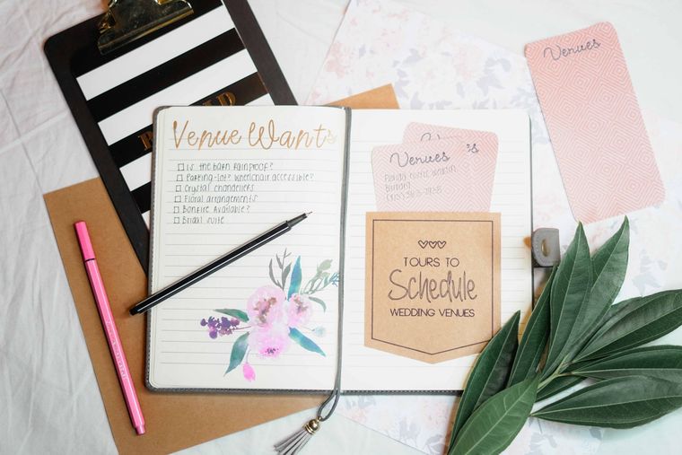 Desk flat lay with pastel stationery, notepad, pink pen, pencil, clipboard, and green leaves on a white surface