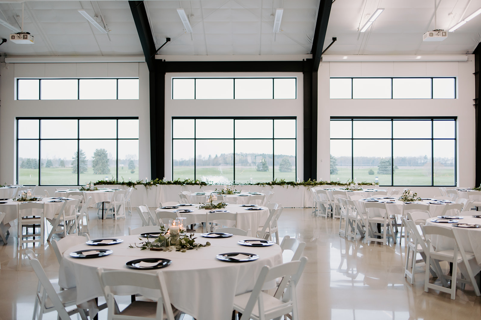 Bright banquet hall with round white tables, chairs, and large windows overlooking a grassy landscape