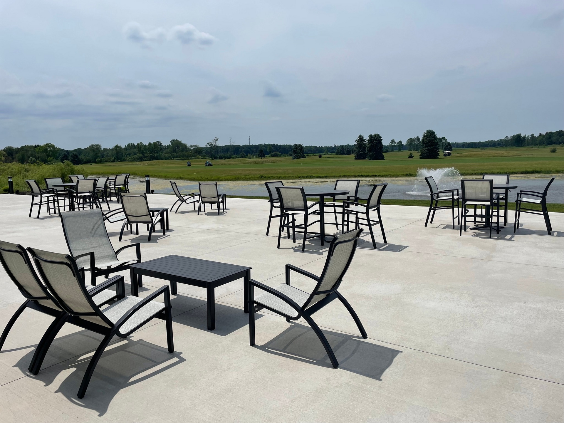 Empty outdoor patio with black chairs and tables overlooking a grassy field under a cloudy sky