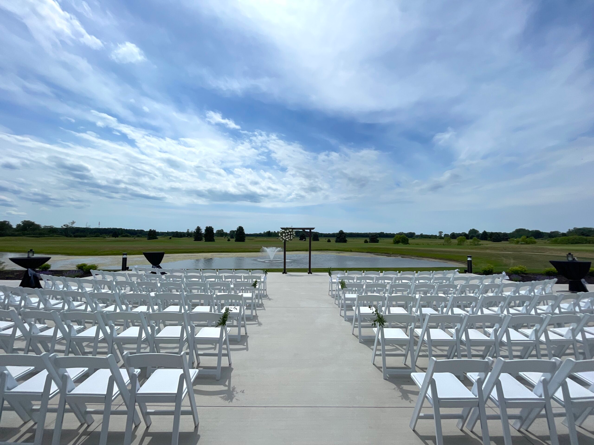 Rows of white chairs facing an outdoor ceremony area under a cloudy blue sky, with open fields beyond.