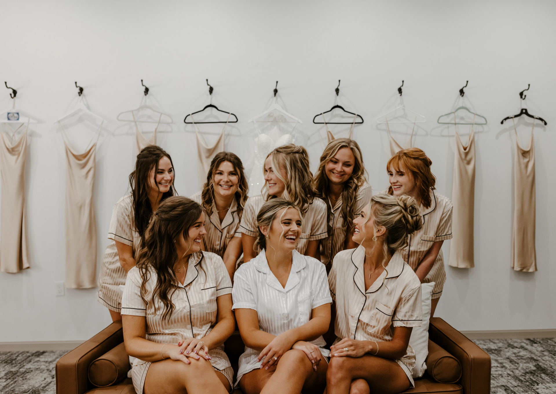 Six women seated and standing in a bridal boutique, smiling among beige dresses on wall hangers