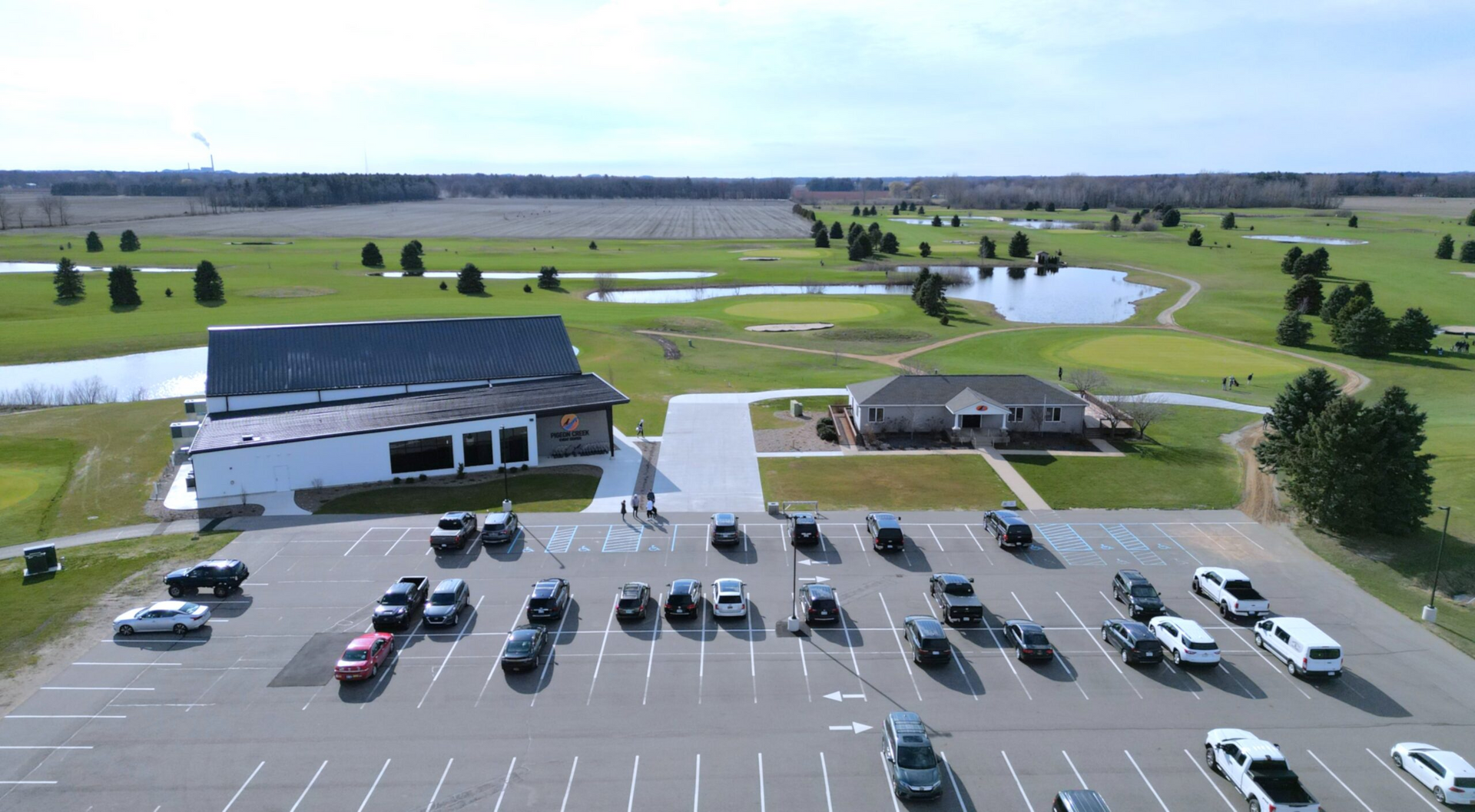 Aerial view of a parking lot beside a building, with a small pond and open fields in the background.