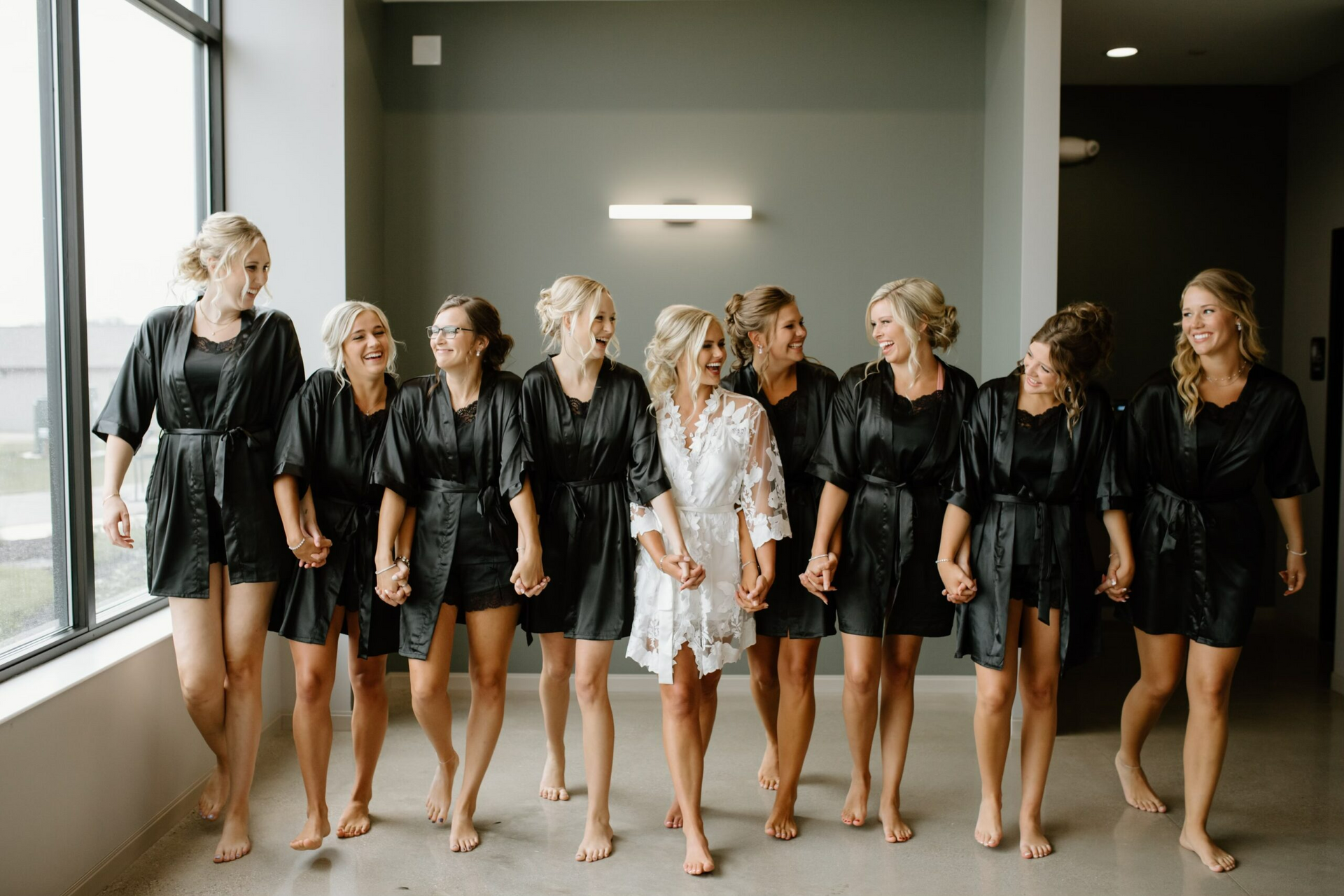Group of women in black robes and one in white robe standing indoors near large windows