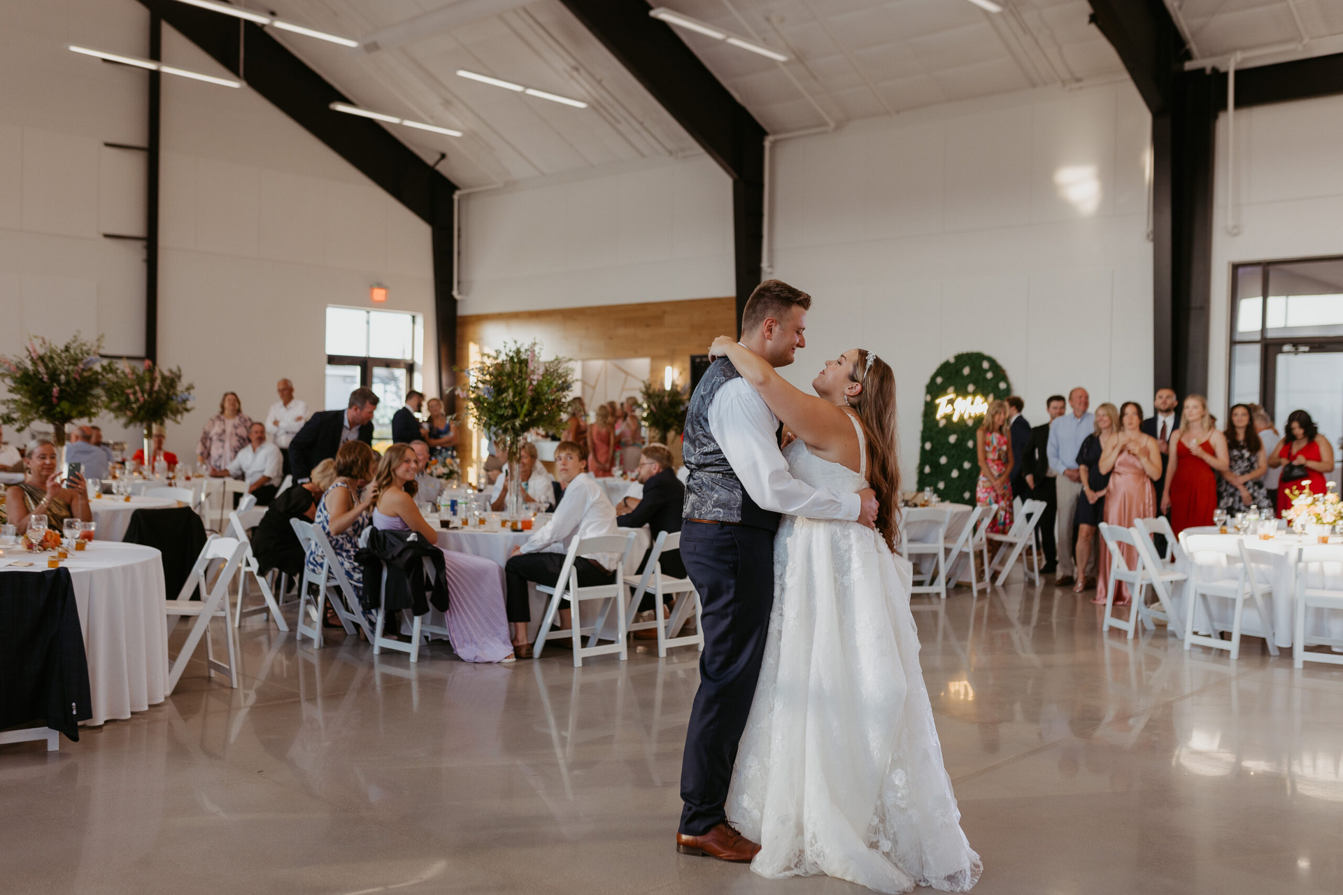 Couple slow dancing at a wedding reception in a bright hall with seated guests and decorated tables