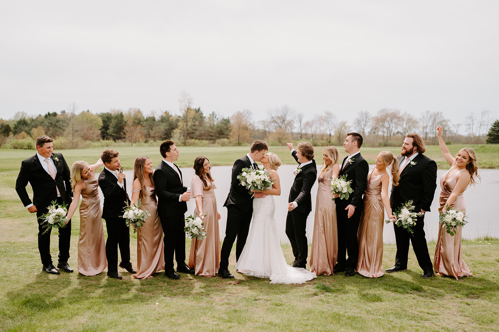 Wedding party standing outdoors in formal attire, holding bouquets on a grassy field.
