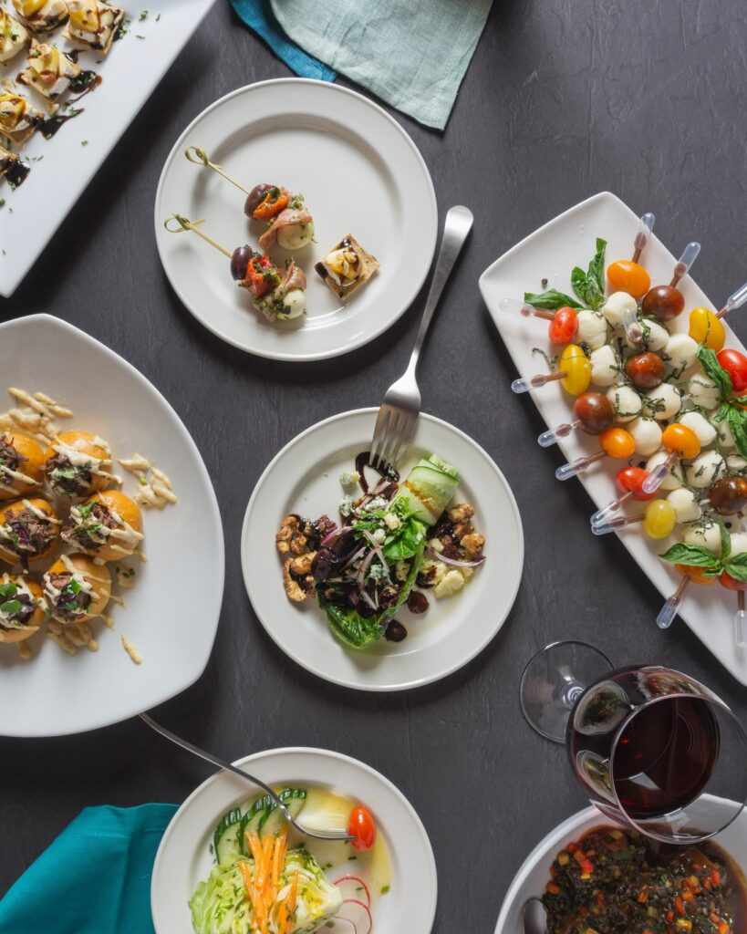 Overhead view of a table with assorted plated appetizers, salads, and drinks arranged on a dark surface