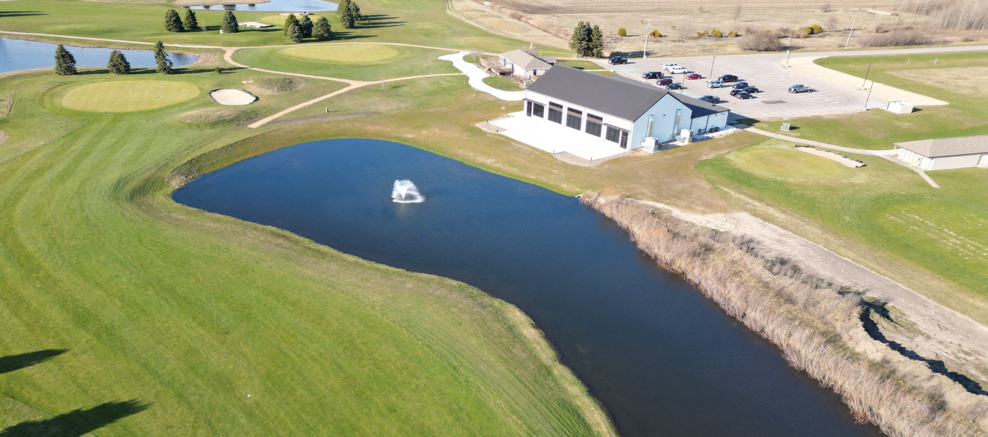 Aerial view of a small pond beside a golf course and buildings, with a fountain in the water