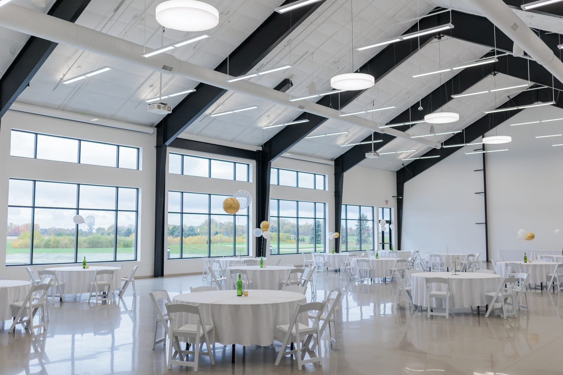 Bright event hall with round tables, white chairs, and large windows overlooking a grassy landscape