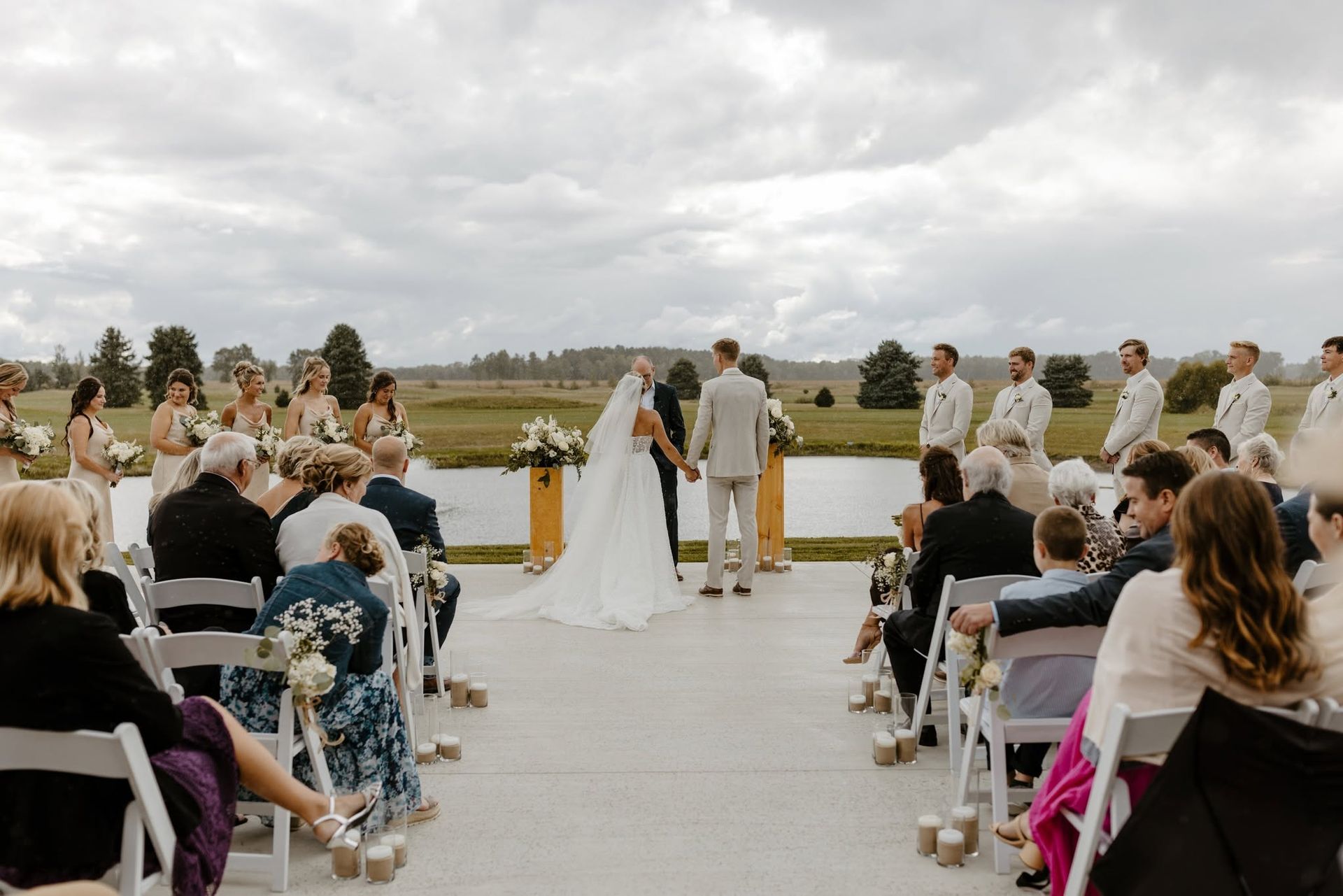 Outdoor wedding ceremony by a lake, with bride and groom at the altar and guests seated on white chairs.