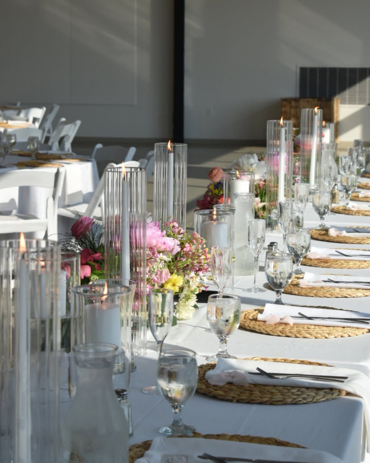 Long banquet table set with white linens, glassware, candles, and pink floral centerpieces by a window