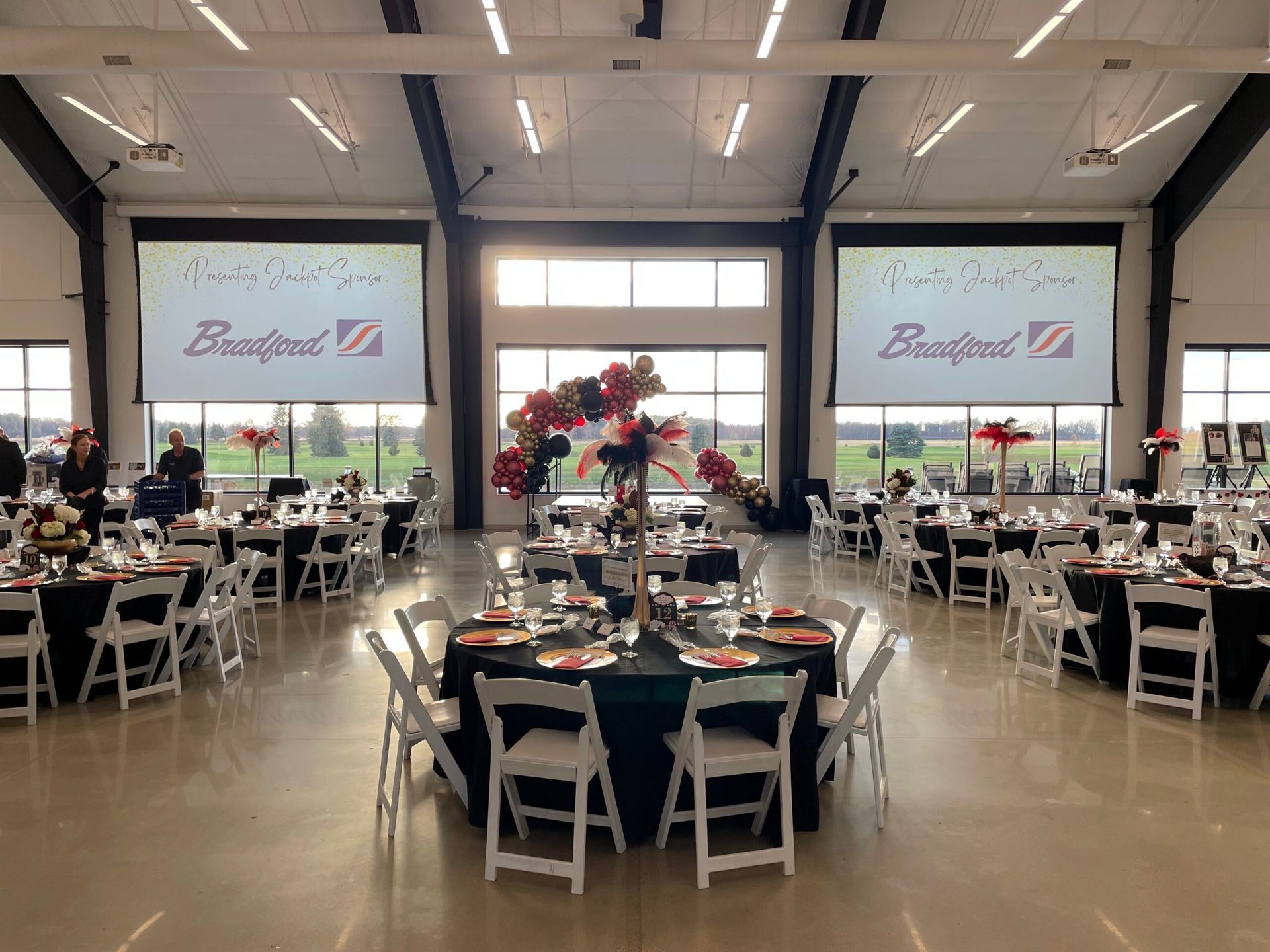 Large banquet hall with round tables, white chairs, and floral centerpieces under bright overhead lights.