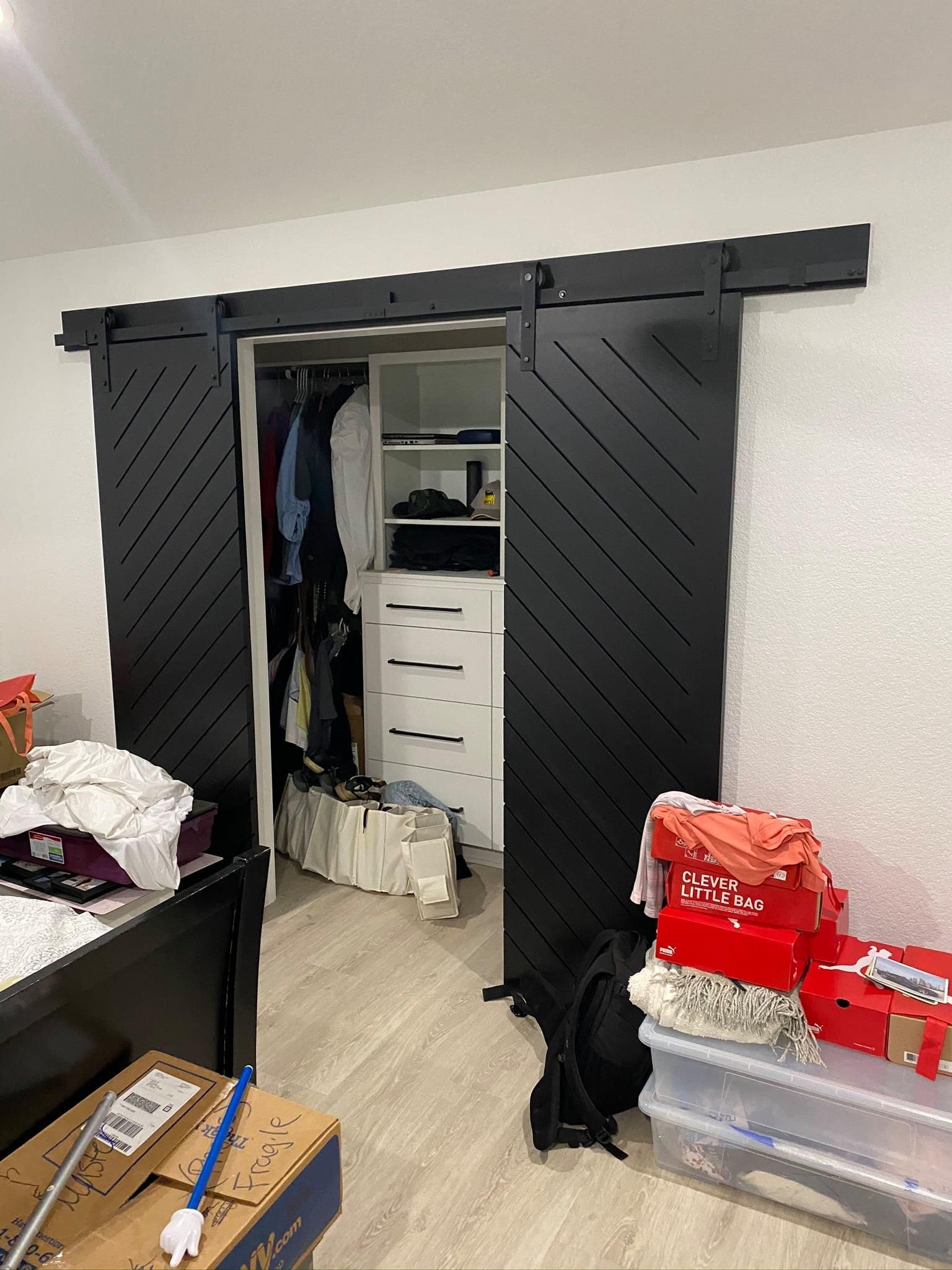 Open closet with sliding barn doors in a cluttered bedroom, showing clothes and white drawers inside.
