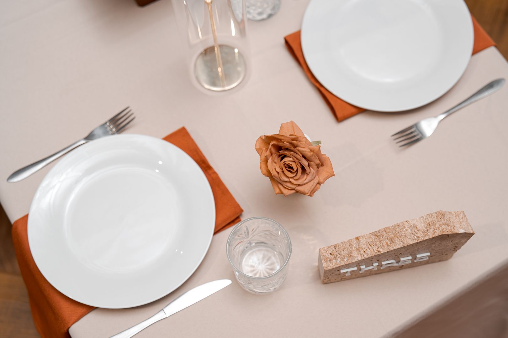 Elegant dining table set with white plates, orange napkins, glasses, a rose centerpiece.
