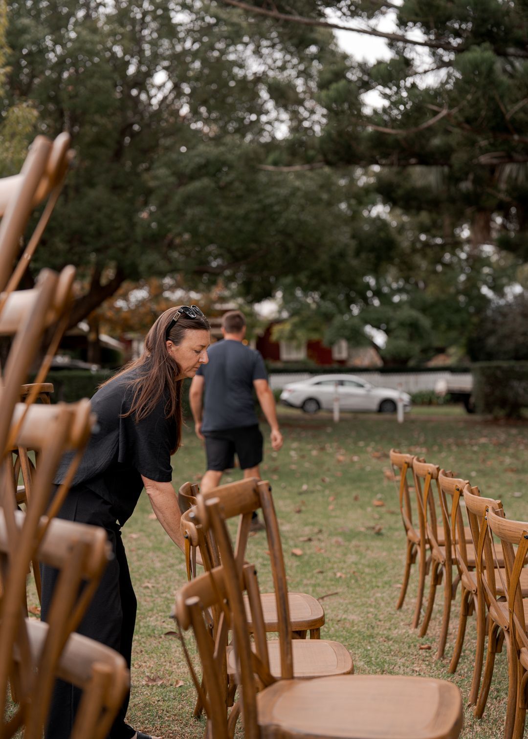 Rows of wooden chairs set up on grass beneath trees, with two people arranging them outdoors.