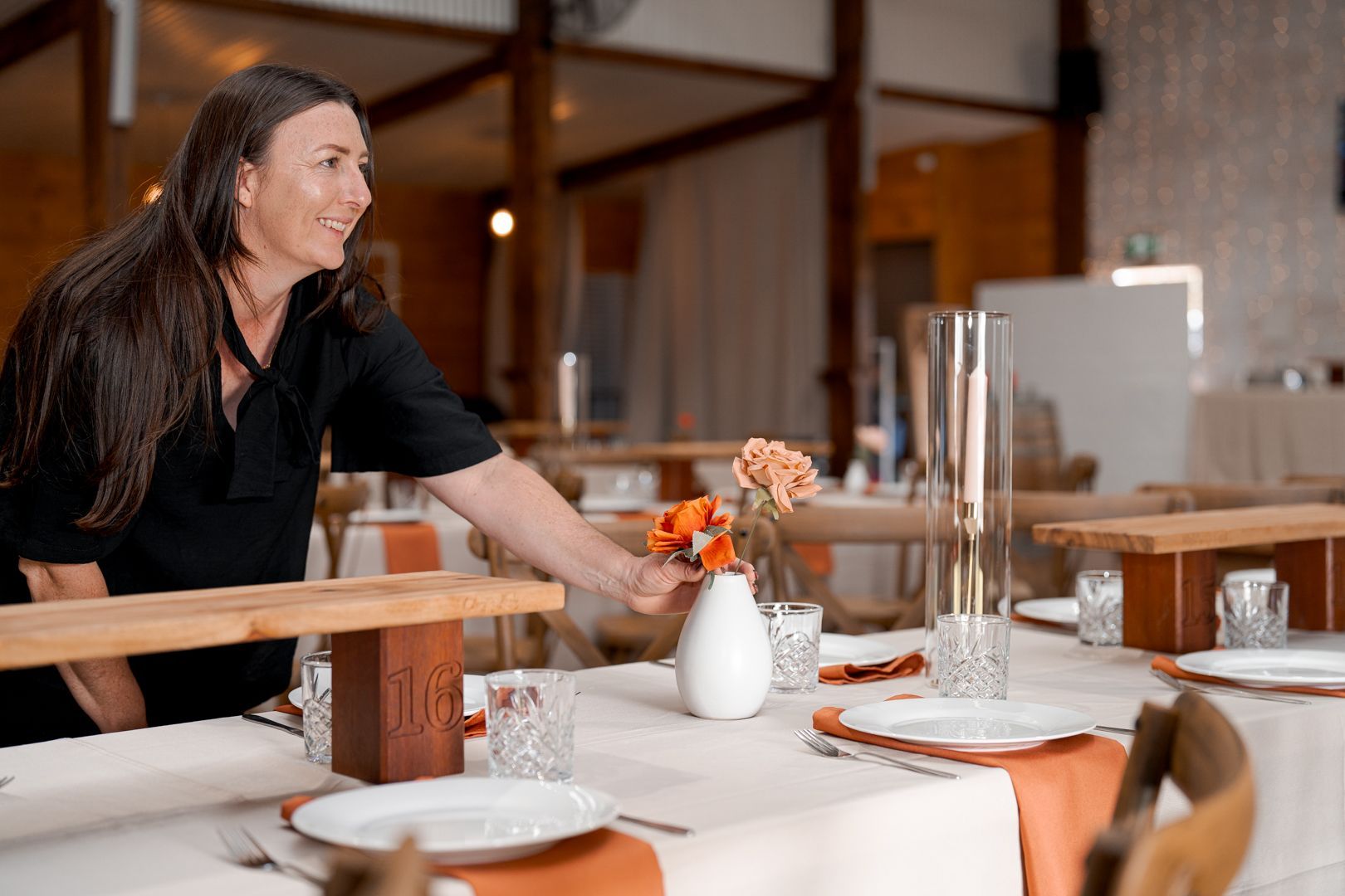 Woman arranging a table with white plates and orange flowers in a rustic venue