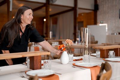 Woman arranging a table with white plates and orange flowers in a rustic venue.