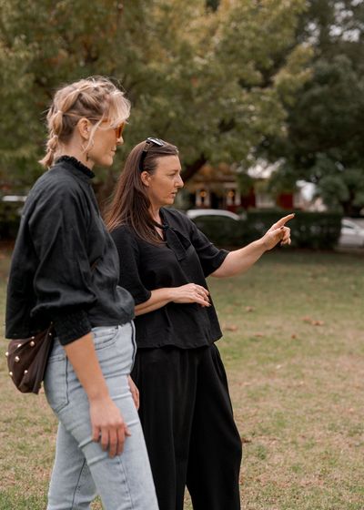Two women standing in a park, one pointing ahead while the other looks on.