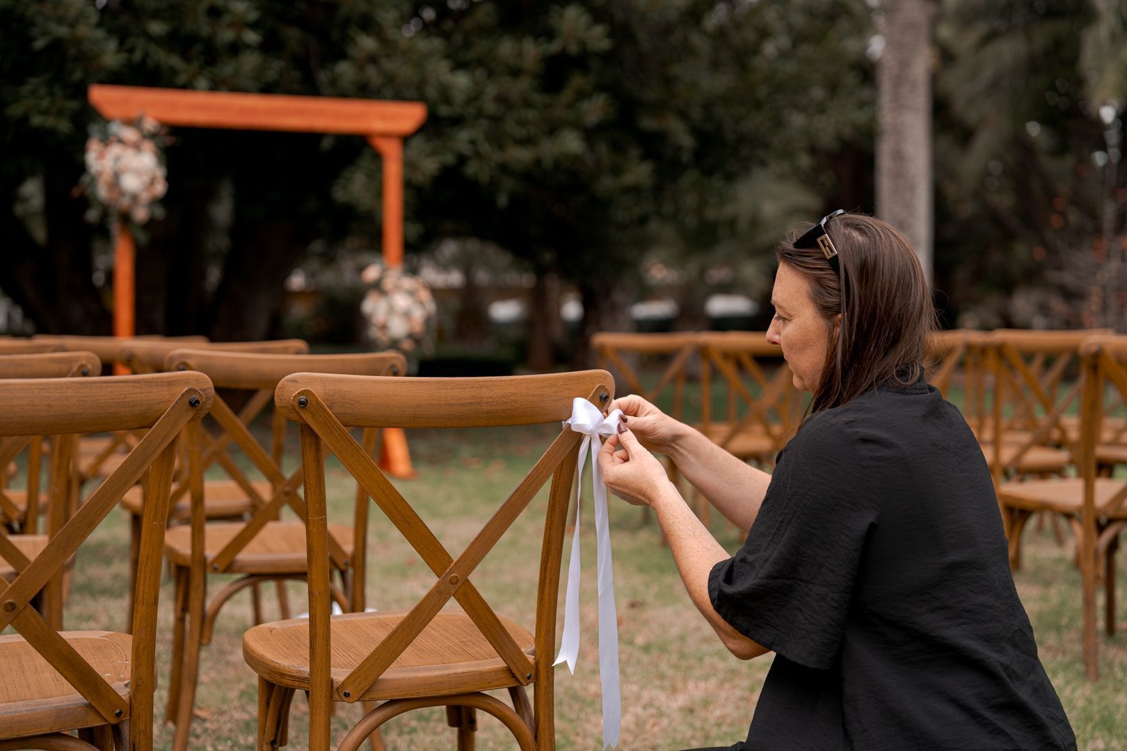Woman tying a white ribbon to a wooden chair in an outdoor ceremony setting
