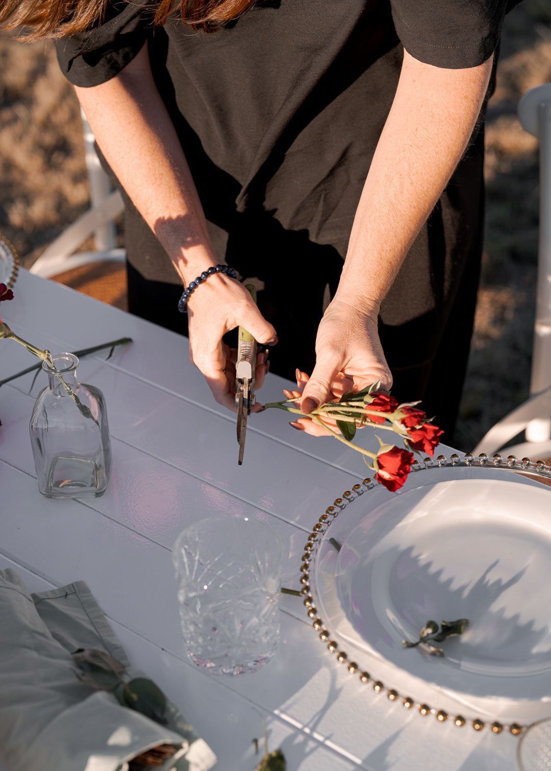 Person arranging red flowers on a table with glassware and a white plate outdoors