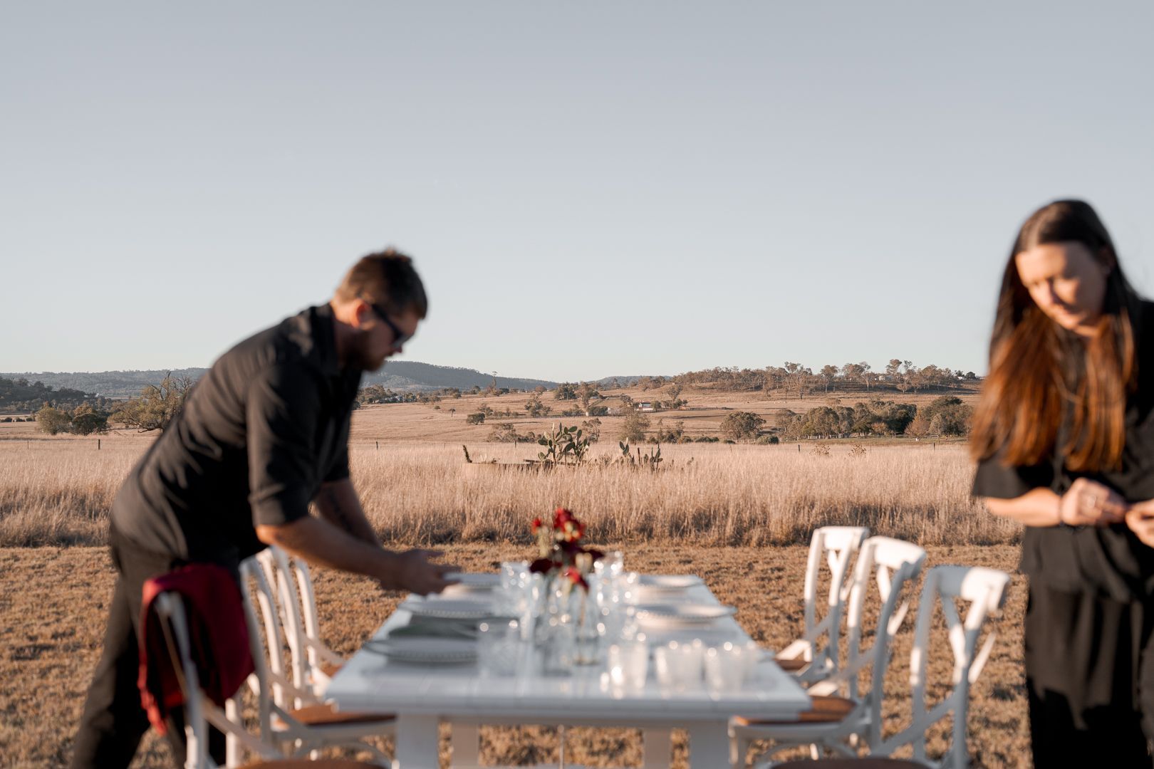 Two people setting a long outdoor dining table in a sunlit field