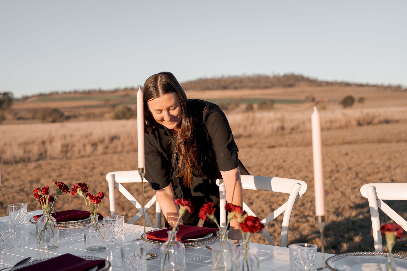Woman arranging red flowers on a white table outdoors in a dry field setting