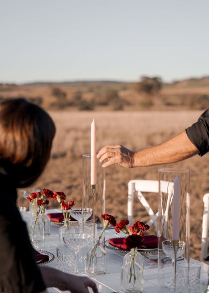 Outdoor table set for a formal dinner, with wine glasses, red flowers, and hands arranging a candle outdoors