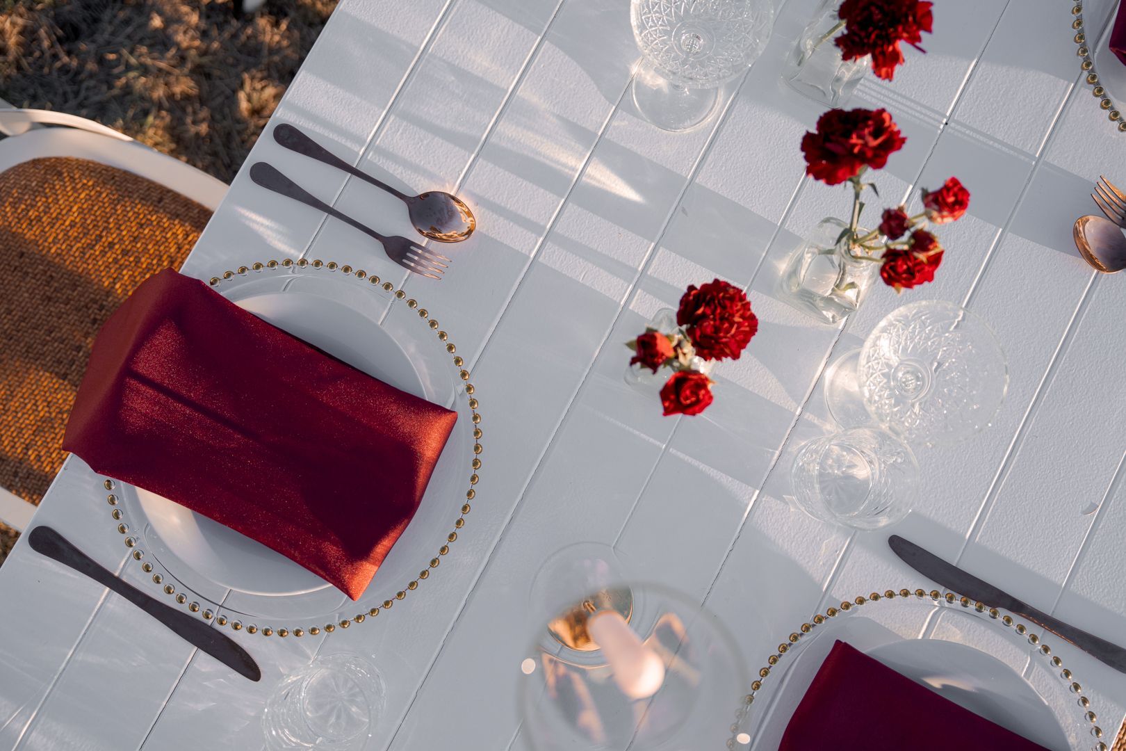 Elegant table setting with burgundy napkins, gold-rimmed plates, candles, and red floral petals on a white tablecloth