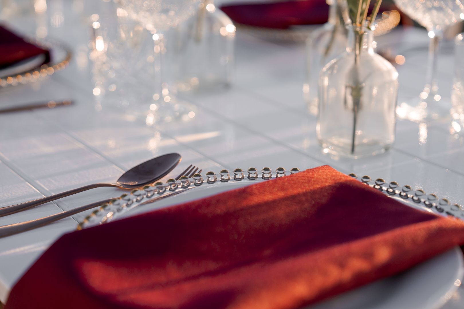 Elegant table setting with red napkins, clear glassware, and small red floral centerpieces.